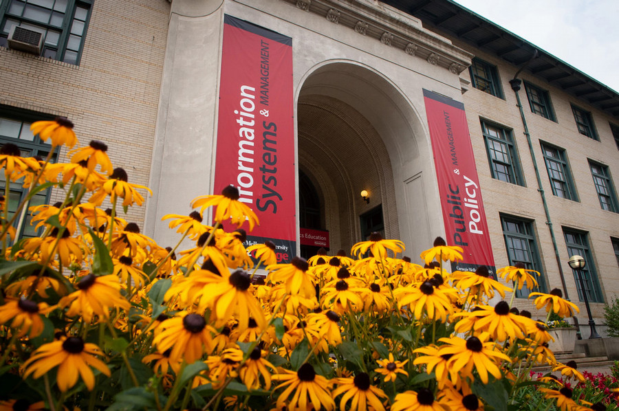 The entrance to the Heinz College of Information Systems and Public Policy at Carnegie Mellon University. The entrance is framed by a large archway with two red banners hanging down the sides, one reading "Information Systems & Management" and the other reading "Public Policy & Management". A bed of yellow flowers sits in front of the entrance.